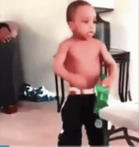 A Shirtless Baby Is Standing In A Living Room Holding A Green Bottle Of Water . GIF