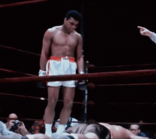 A Shirtless Boxer Stands In The Middle Of A Boxing Ring While A Referee Points At Him GIF