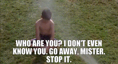 A Shirtless Boy Is Sitting In The Grass With A Sprinkler Spraying Water On Him . GIF