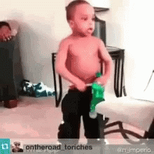 A Shirtless Little Boy Is Standing In A Living Room Holding A Green Toy . GIF