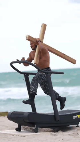 A Shirtless Man Carrying A Wooden Cross On A Treadmill On The Beach GIF