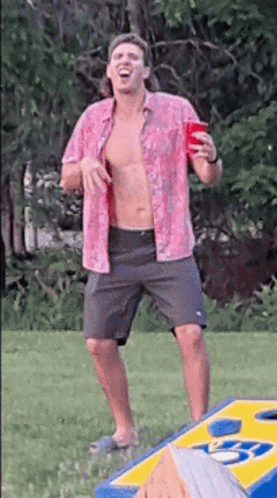A Shirtless Man In A Pink Shirt Is Holding A Red Cup While Standing Next To A Cornhole Board . GIF