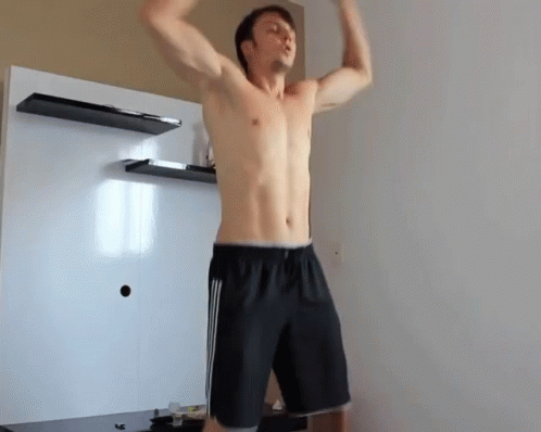 A Shirtless Man In Black Shorts Is Standing In A Room With His Arms In The Air GIF