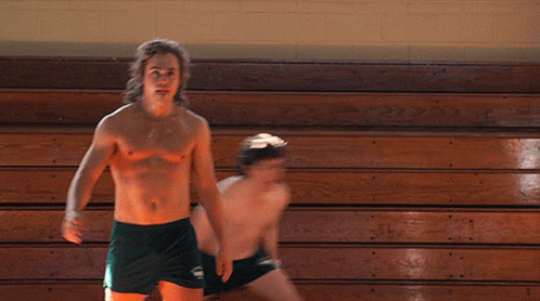 A Shirtless Man In Green Shorts Stands Next To Another Shirtless Man In Black Shorts GIF