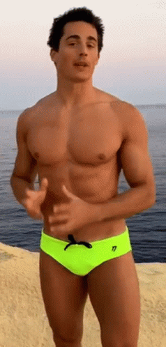 A Shirtless Man In Neon Yellow Swim Trunks Is Standing On The Beach Near The Ocean . GIF