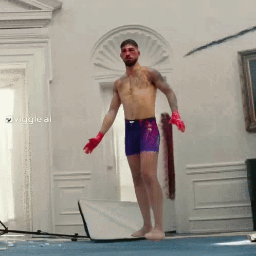 A Shirtless Man In Purple Shorts And Red Gloves Stands On A Blue Mat In A Room GIF