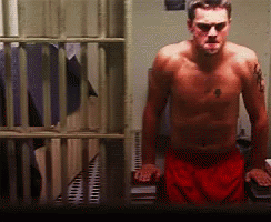 A Shirtless Man In Red Shorts Is Standing In Front Of A Jail Cell GIF