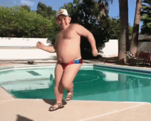 A Shirtless Man In Red Swim Trunks And A Hat Is Running By A Pool GIF