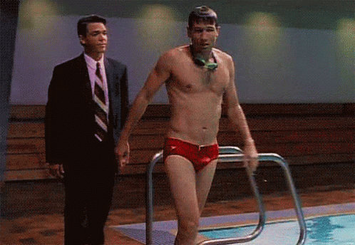 A Shirtless Man In Red Swim Trunks Is Getting Out Of A Swimming Pool While A Man In A Suit Watches . GIF