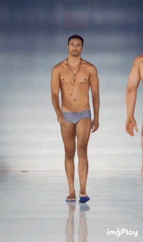 A Shirtless Man In Swim Trunks Walks Down A Runway At A Fashion Show GIF