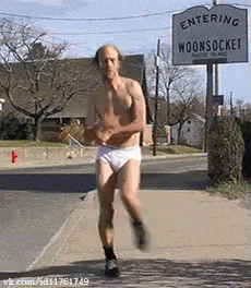 A Shirtless Man In White Underwear Is Running Down A Street In Front Of A Sign That Says Entering Woonsocket GIF