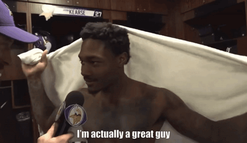 A Shirtless Man Is Being Interviewed In A Locker Room By A Man Wearing A Vikings Hat GIF