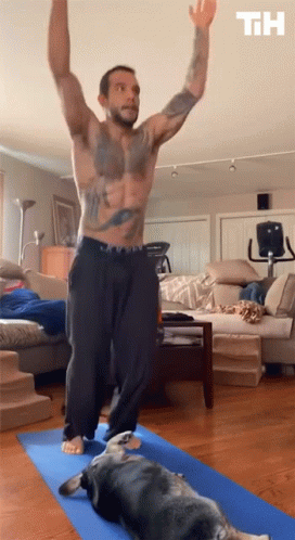 A Shirtless Man Is Doing Yoga In A Living Room With A Dog Laying On A Blue Mat GIF