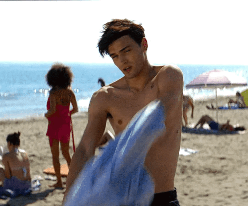 A Shirtless Man Is Holding A Blue Towel On A Beach GIF