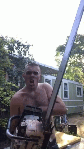 A Shirtless Man Is Holding A Chainsaw In Front Of A House GIF