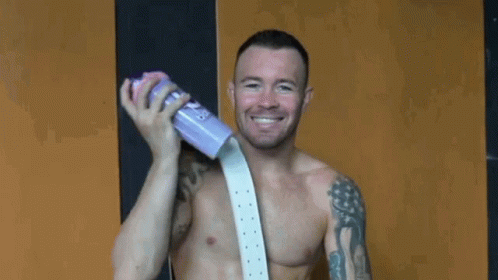 A Shirtless Man Is Holding A Purple Cup And Smiling While Wearing A White Belt . GIF