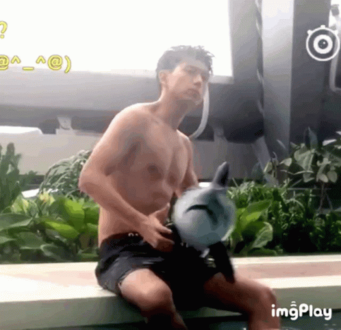 A Shirtless Man Is Holding A Stuffed Dolphin While Sitting Next To A Swimming Pool GIF