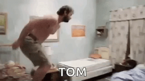 A Shirtless Man Is Jumping On A Bed In A Bedroom With The Word Tom Written On It . GIF