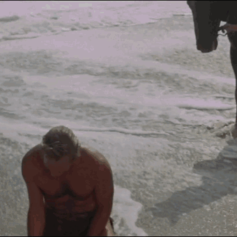 A Shirtless Man Is Kneeling On The Beach Near The Ocean GIF