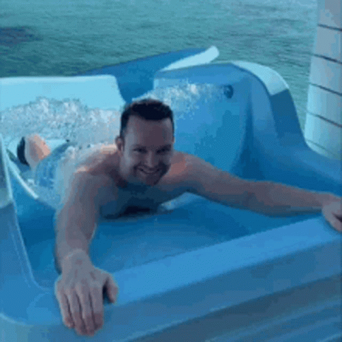 A Shirtless Man Is Laying In A Blue Tub With Water Coming Out Of It . GIF
