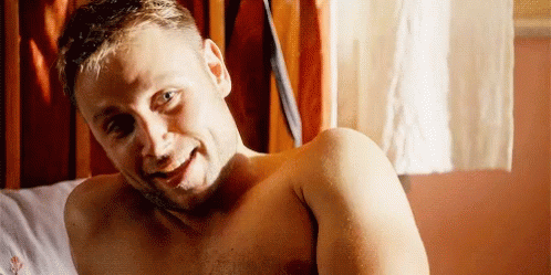 A Shirtless Man Is Laying On A Bed Smiling At The Camera . GIF