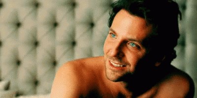 A Shirtless Man Is Laying On A Bed Smiling . GIF