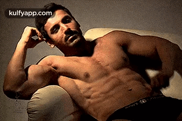 A Shirtless Man Is Laying On A Couch With His Hands On His Head . GIF