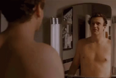 A Shirtless Man Is Looking At His Reflection In A Bathroom Mirror . GIF