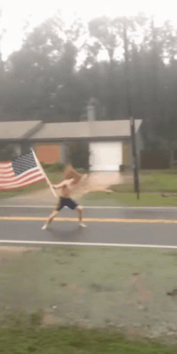 A Shirtless Man Is Running Down A Street With An American Flag In The Background GIF