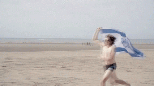 A Shirtless Man Is Running On The Beach Holding A Flag With A Blue And White Star On It GIF