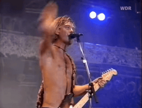 A Shirtless Man Is Singing Into A Microphone While Playing A Guitar GIF