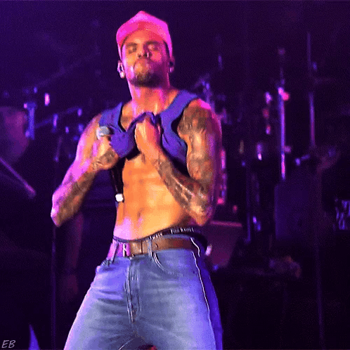 A Shirtless Man Is Singing Into A Microphone While Wearing A Hat GIF