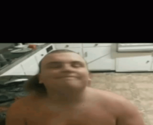 A Shirtless Man Is Sitting In A Kitchen And Smiling At The Camera . GIF