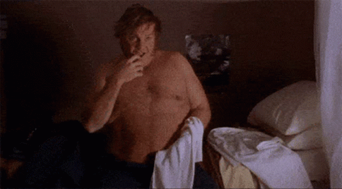 A Shirtless Man Is Sitting On A Bed With A Towel Around His Neck . GIF