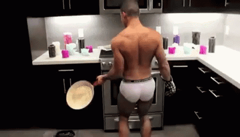 A Shirtless Man Is Standing In A Kitchen Holding A Pan . GIF