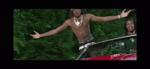 A Shirtless Man Is Standing In Front Of A Car With His Arms Outstretched And A Bandana On His Head . GIF