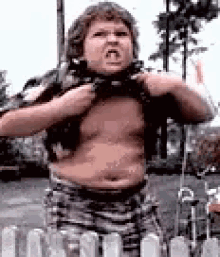 A Shirtless Man Is Standing In Front Of A Fence Holding His Shirt Up . GIF