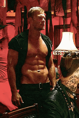 A Shirtless Man Is Standing In Front Of A Lamp With The Letter T On It GIF