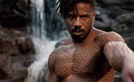 A Shirtless Man Is Standing In Front Of A Waterfall And Looking At The Camera . GIF