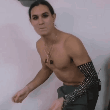 A Shirtless Man Is Standing In Front Of A White Wall Wearing A Necklace And A Bracelet . GIF