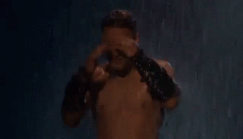 A Shirtless Man Is Standing In The Rain Holding His Head And Wearing Leather Gloves . GIF