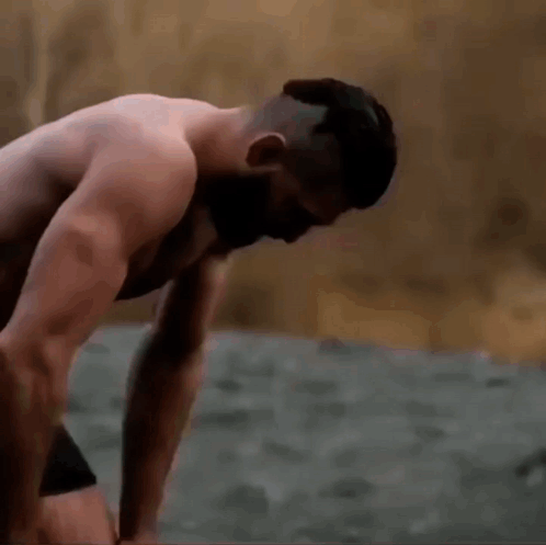 A Shirtless Man Is Standing On A Beach With His Head On His Knees . GIF