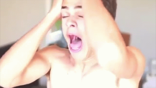 A Shirtless Man Is Yawning With His Mouth Open While Holding His Head In Front Of A Hair Dryer . GIF