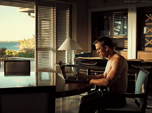 a shirtless man sits at a table with a laptop and a lamp