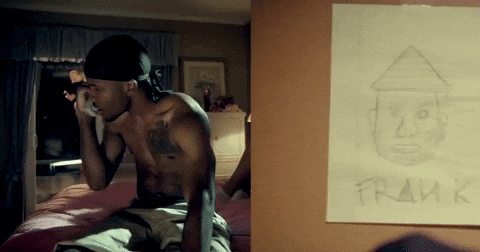 A Shirtless Man Sits On A Bed Next To A Framed Drawing Of Him GIF