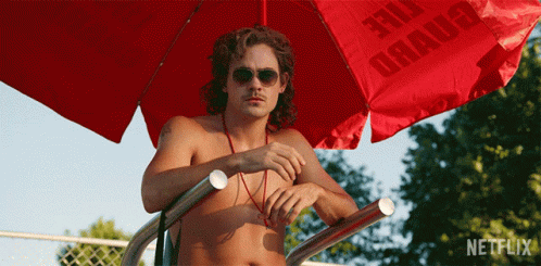 A Shirtless Man Sitting Under A Red Umbrella That Says Netflix On It GIF