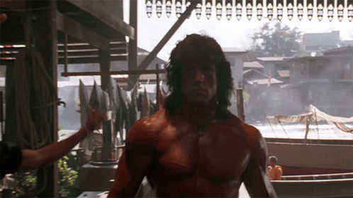 A Shirtless Man Stands In Front Of A Row Of Boats And Buildings GIF