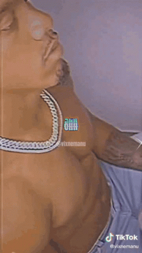 A Shirtless Man Wearing A Chain Around His Neck Is Laying On A Bed . GIF