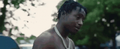 A Shirtless Man Wearing A Chain Around His Neck Is Standing In Front Of Trees . GIF