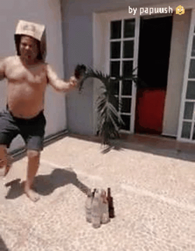 A Shirtless Man Wearing A Paper Hat Is Jumping In The Air While Holding A Palm Tree Branch . GIF